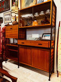 This mid-century modern Ladderax shelving unit features wooden shelves and glass display cabinets framed by black metal supports. A vintage typewriter and ceramic pieces are displayed on the shelves, with framed artwork and other decorative items hanging behind the wall.