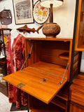 This mid-century modern Ladderax shelving unit features wooden shelves and glass display cabinets framed by black metal supports. A vintage typewriter and ceramic pieces are displayed on the shelves, with framed artwork and other decorative items hanging behind the wall.